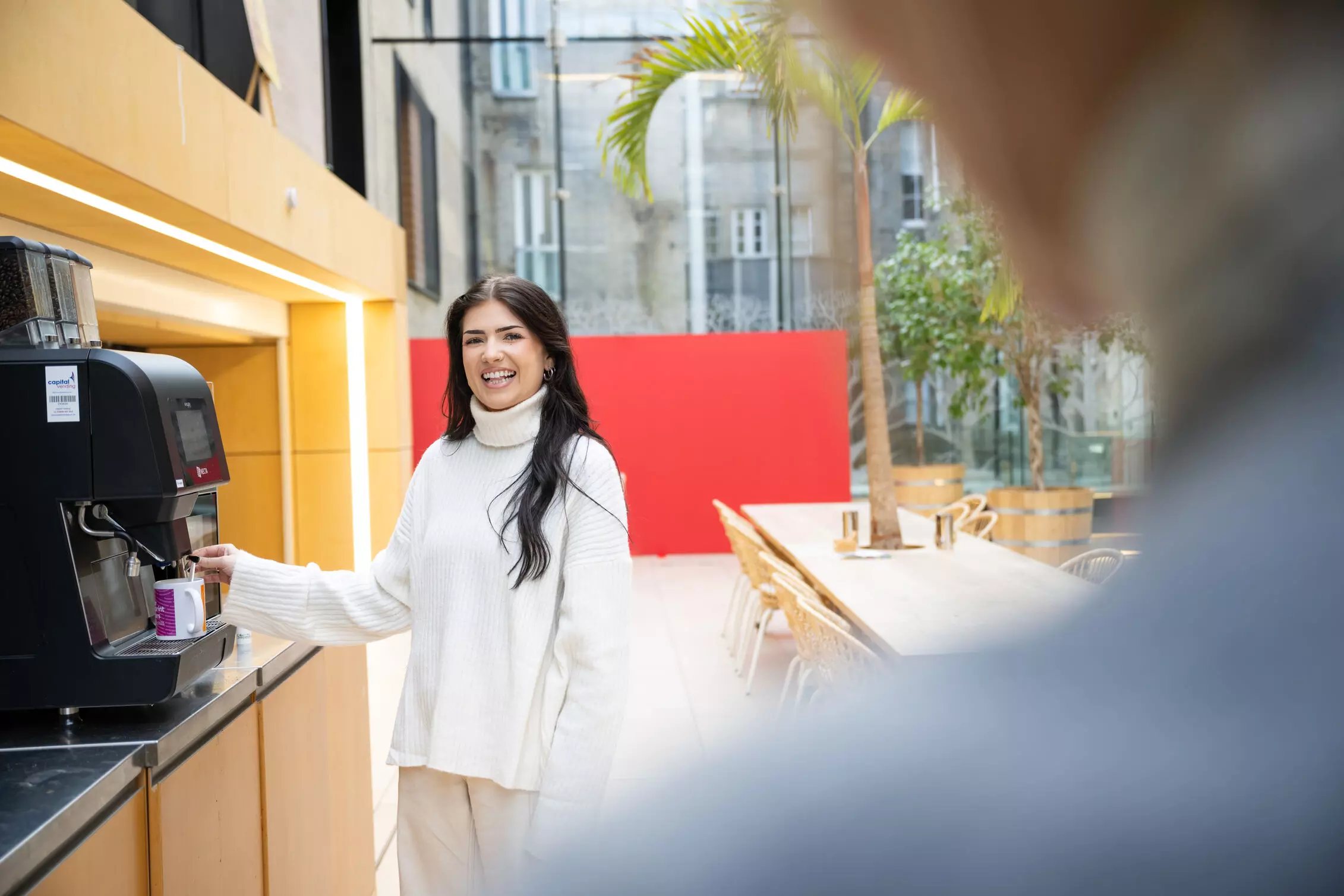 A man and woman wearing headphones are sitting at a desk with a laptop, surrounded by office furniture in an indoor setting.