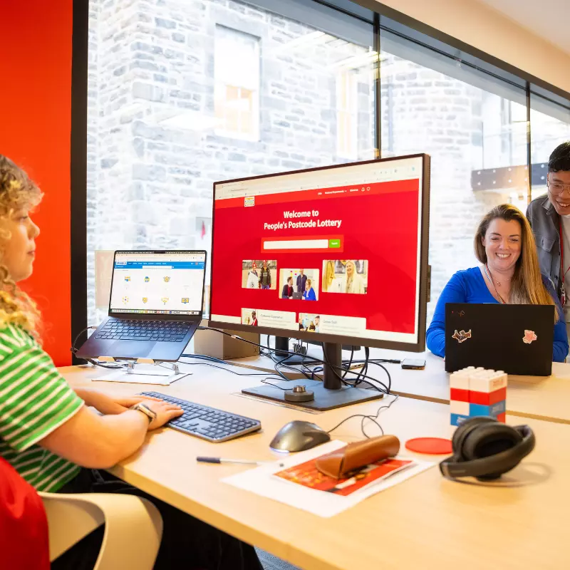 Employee sitting at desk, looking at computer screen which shows our career website.

Across from employee is a further two employees crouched around a laptop screen